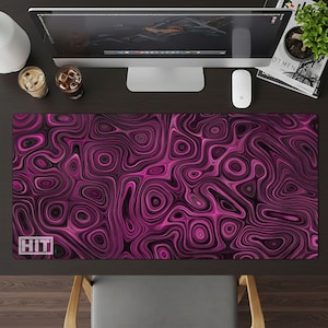 May include: A rectangular desk mat with a swirling, abstract pattern in shades of pink and black. The mat has the word "HIT" in white in the lower left corner. A computer monitor, mouse, and other desk accessories are visible.