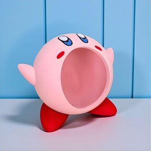 Puede incluir: Un personaje rosa y esférico con una boca grande y abierta, ojos azules y pies rojos. El personaje tiene brazos pequeños y mejillas rojas. La figura está de pie sobre una superficie blanca con un fondo azul claro.