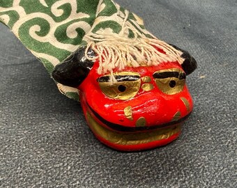 Puede incluir: Una figura de cabeza de león pintada en rojo y oro con detalles en negro y un flequillo de pelo blanco, unida a una bolsa de tela verde y blanca con un patrón de remolinos. El león tiene ojos prominentes, una boca ancha y orejas negras.