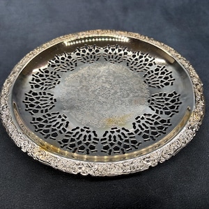 May include: A round silver-toned metal tray featuring a central circular area with an intricate etched pattern, surrounded by a wide border of openwork filigree cutouts and an outer rim decorated with a detailed embossed floral scroll design.