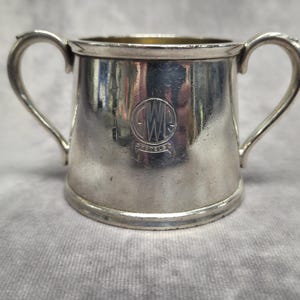 Può includere: Una piccola tazza color argento con due manici curvi. La tazza ha una base arrotondata e un emblema circolare sul davanti. L'emblema sembra avere le lettere "GWR" all'interno di un cerchio. La tazza è probabilmente vintage o antica.