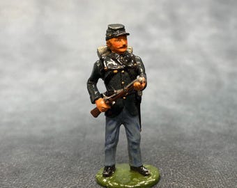 May include: A miniature Civil War soldier figurine. The figure is dressed in a dark blue uniform with a black hat and holds a rifle. The figurine stands on a green base. The soldier has a painted mustache and a stern expression.