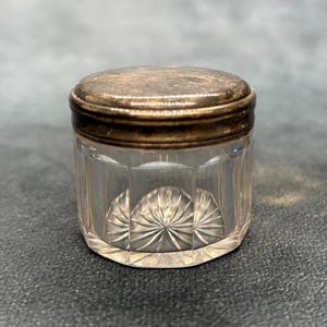 Könnte beinhalten: Ein kleines, klares Glas mit einem silberfarbenen Deckel. Das Glas hat ein geschliffenes Design auf der unteren Hälfte und einen silbernen Rand. Der Deckel ist angelaufen, was ihm ein Vintage-Aussehen verleiht. Das Glas dient wahrscheinlich zur Aufbewahrung kleiner Gegenstände.