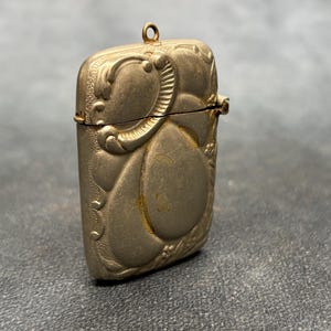 Pode incluir: Uma caixa de fósforos retangular de metal com acabamento dourado fosco, apresentando padrões orgânicos fluidos em relevo e um pequeno anel circular no topo. O estojo possui uma dobradiça horizontal no meio para abrir a parte superior.