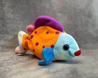 May include: A plush fish toy featuring a bright orange body with purple polka dots, a light blue head, a purple dorsal fin, a blue pectoral fin, a white tail, and red lips. The fish has large black plastic eyes and a soft, fuzzy texture.