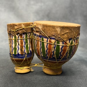 May include: A double-headed drum instrument featuring two connected ceramic bodies with tan, green, blue, and brown vertical stripes and wavy lines. The drum heads are made of light brown animal skin, secured with a gold-colored string netting.