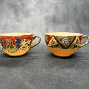 Puede incluir: Dos tazas de té antiguas con diseños florales y geométricos. Una taza presenta un borde rojo y naranja con un motivo floral, mientras que la otra tiene un patrón geométrico con detalles rojos, verdes y azules. Ambas tienen asas doradas.