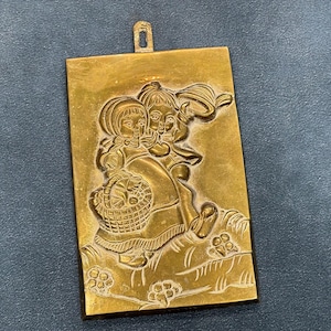 May include: A rectangular, gold-colored pendant with a detailed relief of two children. One child carries a basket, and both are depicted on a grassy hill with floral accents. The pendant has a small loop at the top for hanging.