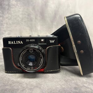 Puede incluir: Cámara Halina 35-600 negra con funda de cuero. La cámara tiene una lente plateada y el nombre "HALINA" está impreso en blanco. La lente está marcada "ANASTIGMAT" y "F=40mm". La funda de cuero negra tiene un borde plateado.