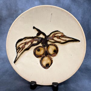 May include: A round, off-white ceramic plate featuring a hand-painted, dark brown botanical illustration of three rounded fruits clustered together with two elongated leaves extending outward, rendered in a rustic, textured glaze finish.