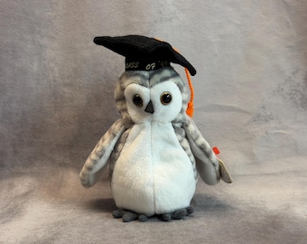 May include: A plush owl toy with gray and white fur, large dark eyes, and a black graduation cap featuring the text "CLASS OF '99" in white embroidery and an attached orange tassel hanging from the side of the cap.