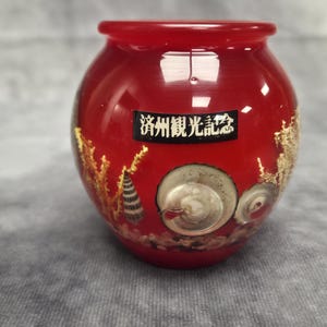 May include: A red, round decorative vase with a glossy finish. The vase features gold accents and illustrations of seashells. A rectangular label with Japanese text is affixed to the front. This item is a souvenir.