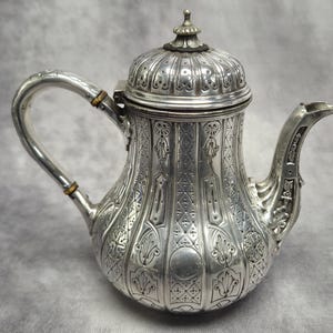 Puede incluir: Tetera plateada ornamentada con asa curva y boquilla. La tetera presenta un diseño detallado en relieve que cubre el cuerpo y la tapa. La tapa está rematada con un pequeño adorno decorativo. Probablemente vintage o antigua.