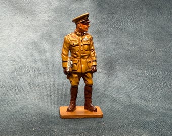 May include: A hand-painted metal figurine of a military officer standing at attention. The figure wears a tan uniform with a matching peaked cap, dark brown tall boots, and holds a small white object in the right hand while standing on a base.