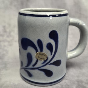 May include: A stoneware beer stein with a light gray base and a dark blue floral design. The mug has a handle and a dark blue band around the top and bottom. A small sticker is affixed to the front.