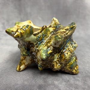 May include: A ceramic seashell-shaped planter with a textured surface. The planter features a blend of green, brown, and blue hues, with a speckled, glazed finish. It has a small opening at the top and is supported by three small feet.