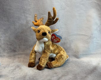 May include: A plush deer toy with brown and tan textured fur, a white chest, and dark brown hooves. The deer features large antlers with a patterned surface and a heart-shaped red and yellow Ty tag attached to its ear. The toy has black eyes.