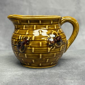 May include: A small, brown ceramic creamer with a basket weave design. The creamer has a handle and a spout for pouring. Two floral accents are visible on the front. The creamer is a warm, earthy tone.