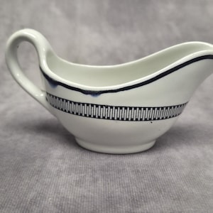 Puede incluir: Salsera de cerámica blanca con asa y boquilla. El recipiente está decorado con un borde azul oscuro a lo largo del borde y una banda de líneas verticales en el medio. La salsera está sobre una superficie gris.