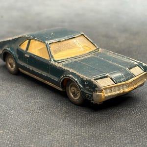 May include: A vintage dark green die-cast toy car with a tan interior and visible paint wear along the edges. The model features a front grille, silver-toned wheels, and a clear windshield, showing signs of age and play with chipped paint.