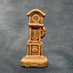 May include: A miniature tan ceramic grandfather clock figurine with a glossy finish. The clock face is at the top, with a central panel and a rectangular base. The word WADE is embossed in capital letters on the front of the base of the figurine.