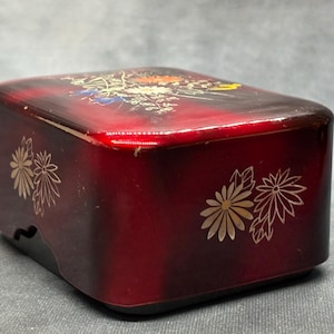 Op de afbeelding: Een rechthoekige, rood gelakte doos met gouden bloemmotieven. De bovenkant heeft een gedetailleerd bloemen- en bladerpatroon in meerdere kleuren. De zijkanten zijn versierd met gouden bloem- en bladmotieven. De doos heeft een glanzende afwerking.
