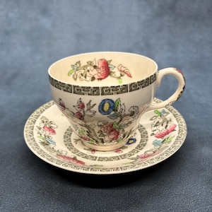 Puede incluir: Un juego de taza y platillo blancos con un diseño floral. La taza y el platillo presentan flores rosas, azules y verdes, con un borde geométrico negro y gris. El juego es de porcelana.