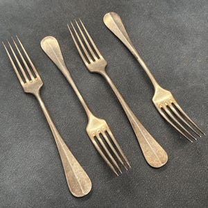 Pode incluir: Um conjunto de quatro garfos de metal prateado com acabamento mate. Cada garfo apresenta um cabo longo e fino que alarga ligeiramente na extremidade e quatro dentes finos e pontiagudos. Os garfos estão dispostos num padrão diagonal.