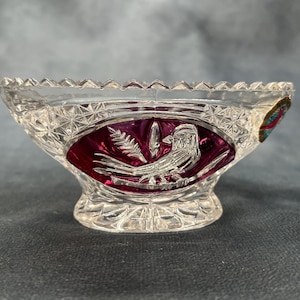 Può includere: Una ciotola trasparente in vetro tagliato con un disegno di uccello e foglie su sfondo bordeaux. La ciotola ha un bordo smerlato e un motivo a stella. Un adesivo colorato è sul lato.