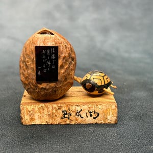 May include: A decorative figurine featuring a brown walnut shell with a black rectangular plaque displaying white Japanese text, positioned next to a small painted turtle on a rectangular wooden base with black Japanese characters inscribed on the front.