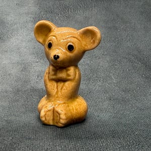 Könnte beinhalten: Eine kleine, glänzende, hellbraune Keramikfigur einer Maus in sitzender Position. Die Maus hat große, abgerundete Ohren, dunkel bemalte Augen, eine kleine schwarze Nase und eine texturierte Oberfläche mit subtilen Variationen in der Glasur.