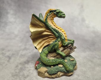 Puede incluir: Figura detallada de un dragón verde y amarillo posado sobre una formación rocosa. El dragón tiene grandes alas color canela y un cuerpo largo y serpentino. La base está detallada con musgo y pequeños detalles rojos.