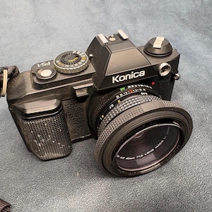 May include: A black Konica FS-1 35mm film camera featuring a textured grip, a top-mounted shutter speed dial, and a lens with text reading Konica Hexanon AR 40mm F1.8 LENS MADE IN JAPAN. The camera includes a black fabric neck strap attached.