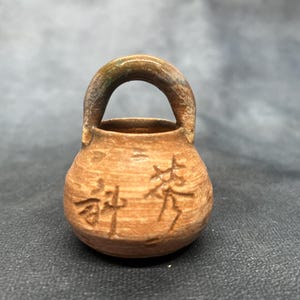 Puede incluir: Una pequeña olla de cerámica hecha a mano con un asa. La olla es de color marrón cálido con una superficie ligeramente texturizada. Dos caracteres chinos están tallados en la parte delantera. El asa tiene un degradado verde y marrón.