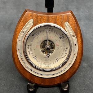 Puede incluir: Un barómetro vintage con un marco de madera en forma de herradura. La esfera plateada indica la presión con palabras como "Tormentoso" y "Bueno". El barómetro tiene una aguja y está montado sobre un soporte negro.