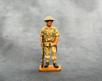 May include: A hand-painted miniature soldier figurine wearing a tan uniform, a matching helmet, and tactical gear. The figure stands on a rectangular base and holds a rifle in one hand, with detailed shading on the clothing and equipment.