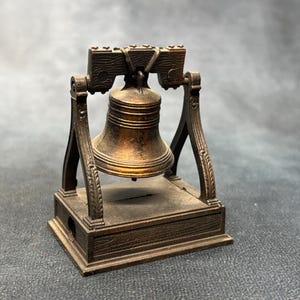 May include: A miniature bronze-toned metal bell suspended within a decorative frame. The bell hangs from a horizontal bar supported by two arched side pillars, all mounted on a rectangular base with a wood-grain texture and a visible horizontal slot.