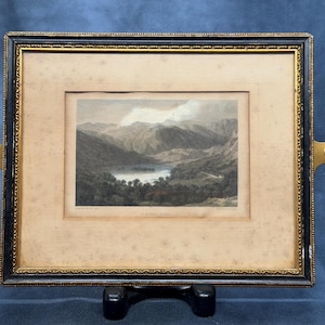 Puede incluir: Una impresión de paisaje enmarcada titulada "Buttermere" que muestra un lago de montaña con un paspartú beige, un marco decorativo negro y dorado, y dos pestañas de montaje laterales de latón. La imagen muestra un valle con colinas y agua.