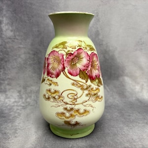 Puede incluir: Un jarrón de cerámica vintage con base y borde verde claro, y cuerpo color crema. El jarrón está adornado con diseños florales rosas y patrones dorados en forma de nube. El jarrón mide aproximadamente 20 cm de alto.