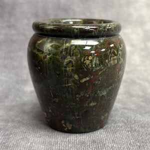 Könnte beinhalten: Eine kleine, runde, dekorative Vase mit einem dunkelgrünen und kastanienbraunen, melierten Muster. Die Vase hat einen leicht ausgestellten Rand und einen abgerundeten Körper. Die Steinvase ist ein einzigartiger Einrichtungsgegenstand.