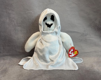 May include: A white plush ghost toy featuring a hooded cape, black embroidered eyes and a jagged mouth. The stuffed figure has a soft, shimmering fabric texture and includes a red heart-shaped Ty tag attached to its side with the letters ty printed.