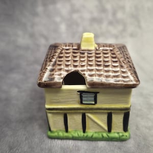 May include: A miniature ceramic house with a brown tiled roof, yellow walls, and a green base. The house has a chimney and a small window. The roof has a hole in it. The house is a decorative item.
