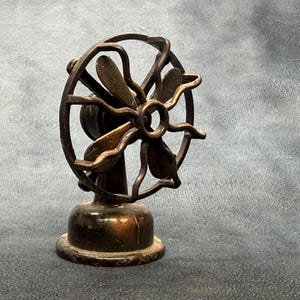 May include: A small, vintage-style desk fan made of dark, weathered bronze-toned metal. It features a circular protective cage with a wavy, decorative frame and four stationary blades centred within the structure, all mounted on a sturdy, round base.