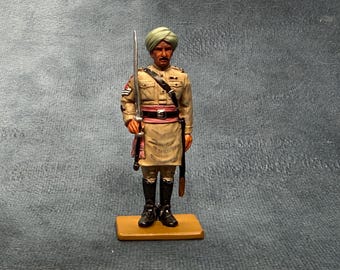 May include: A hand-painted metal figurine of a soldier wearing a tan uniform, a light green turban, and black boots. The soldier stands on a rectangular base, holding a silver-colored sword upright in the right hand and wearing a dark belt.