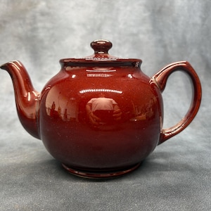 Op de afbeelding: Een glanzende, diep mahoniebruine keramische theepot met een rond lichaam, een gebogen tuit en een bijpassend deksel met een kleine knopgreep. De theepot heeft een glad, reflecterend oppervlak en een stevig handvat aan de rechterkant.