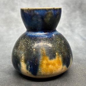Könnte beinhalten: Eine handgefertigte Keramikvase mit einer einzigartigen Form und einer auffälligen Farbpalette. Die Vase hat einen dunkelblauen oberen Teil und einen gesprenkelten, strukturierten Körper mit Schattierungen von Schwarz, Gelb und Weiß. Die Vase ist etwa 13 cm hoch.