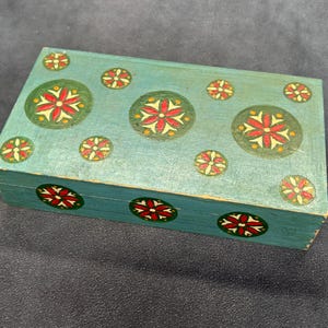 Puede incluir: Una caja rectangular de madera pintada en azul claro, con una serie de motivos circulares en la parte superior y frontal. Cada círculo contiene un diseño floral rojo y amarillo sobre un fondo de color verde oscuro.