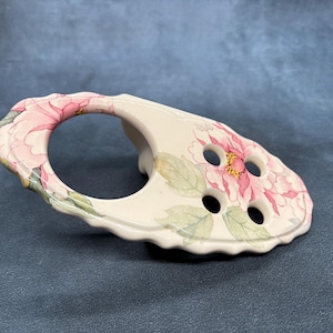 Może przedstawiać: Biały ceramiczny uchwyt na szczoteczki do zębów z kwiatowym wzorem przedstawiającym różowe piwonie i zielone liście. Uchwyt ma cztery okrągłe otwory na szczoteczki do zębów i zakrzywiony uchwyt.