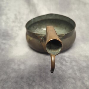 Könnte beinhalten: Ein kleines, antikes Kupfergefäß mit Ausguss und Henkel. Der schalenförmige Körper hat eine dunkle, gealterte Patina, und der Ausguss ist von hellerer Kupferfarbe. Der Henkel ist nach unten gebogen.