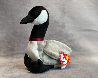May include: A plush Canada goose toy with a black head and neck, a white cheek patch, and a light gray body. The goose features a dark red ribbon around its neck and a heart-shaped red and yellow Ty tag attached to its side wing.